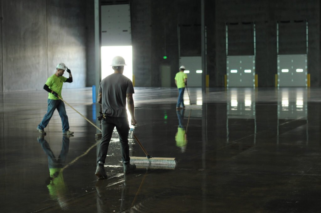 Three concrete flooring applicators with push brooms brooming the Ashford Formula liquid densifier on the floor surface in a newly constructed warehouse.