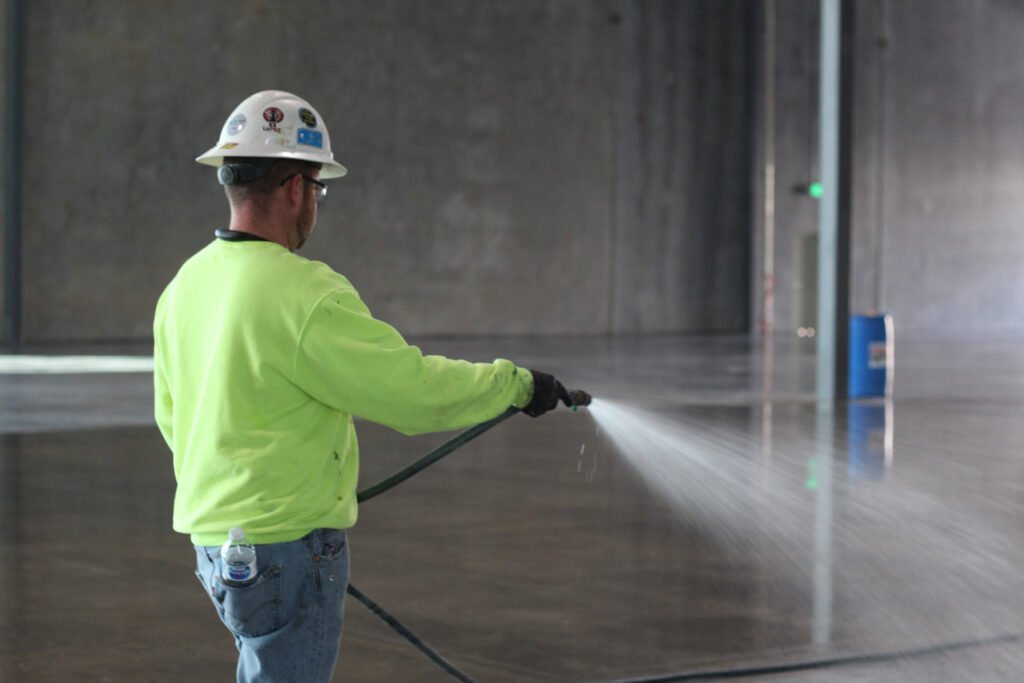 Concrete flooring applicator wearing fluorescent yellow sweater, white hard hat, and safety goggles holding a hose and spraying Ashford Formula densifier on the concrete floor; blue Ashford Formula drum out of focus in the background.
